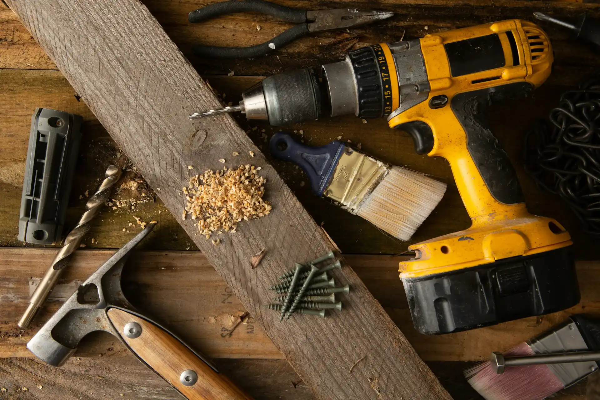 Hand and power tools on a wooden workbench: a cordless drill, a hammer, pliers, drill bits and screws.
